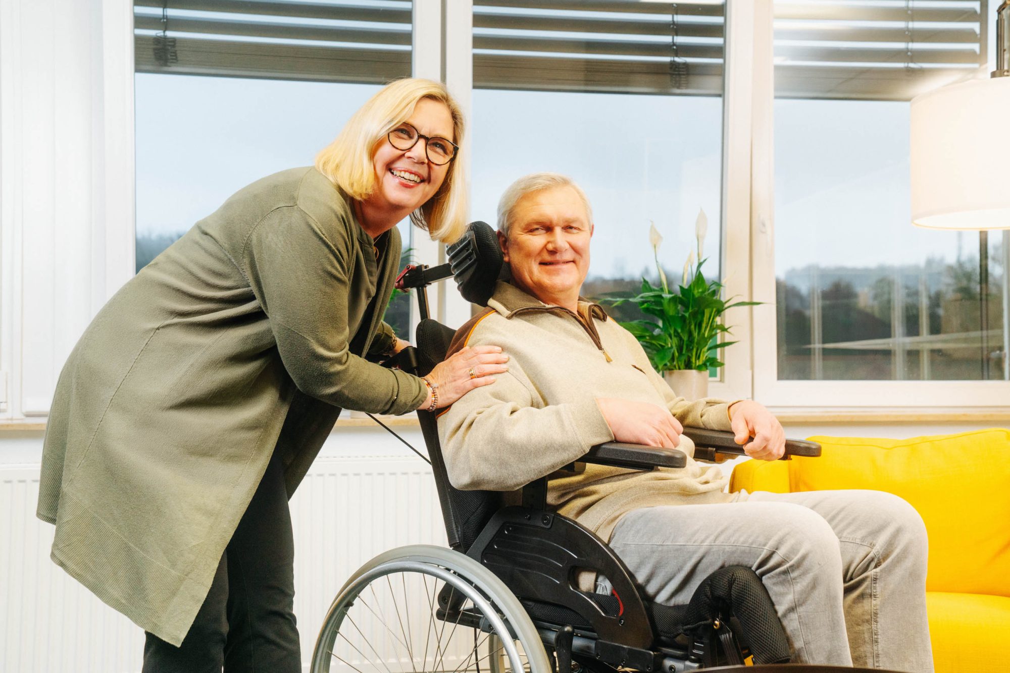 Pflegende Angehörige steht lächelnd neben einem älteren Mann im Rollstuhl