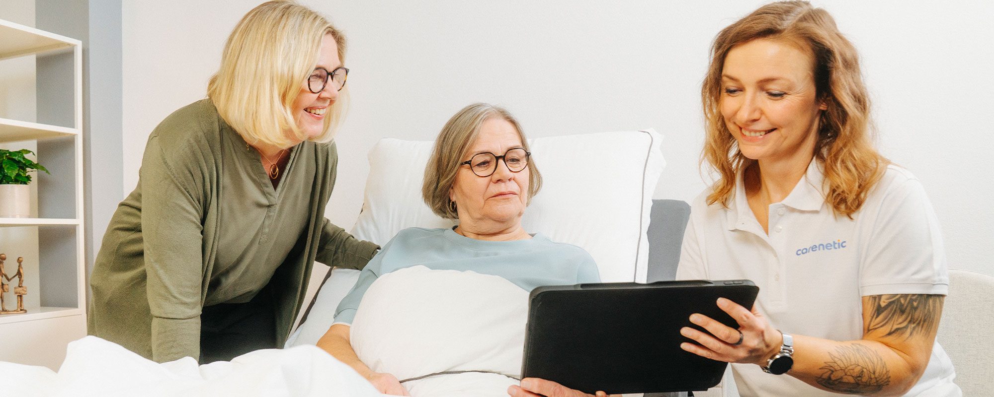 Carenetic Mitarbeiterin zeigt einer Patientin im Pflegebett und ihrer Angehörigen Informationen auf einem Tablet