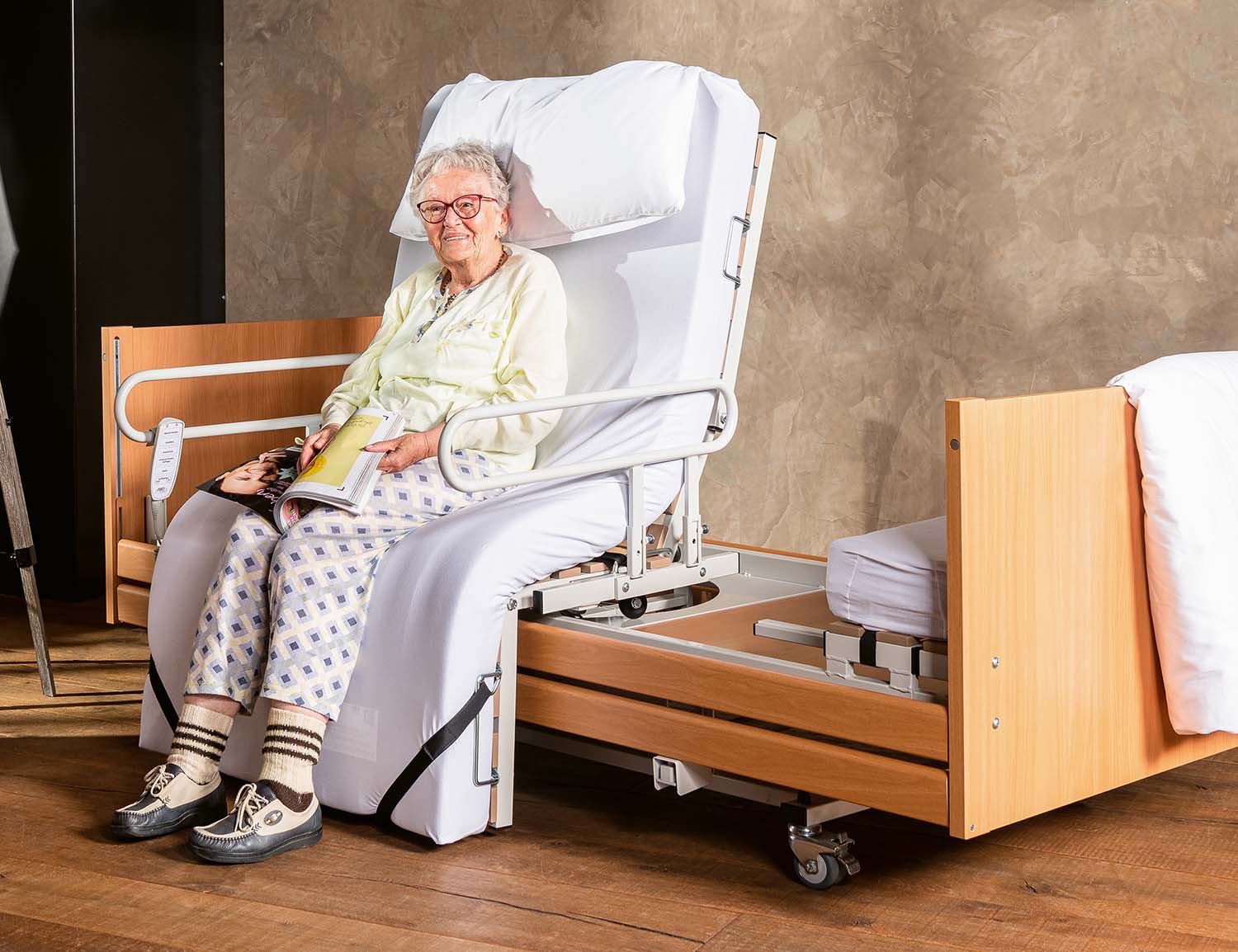 Elderly woman sitting in a specialist bed with the stand-up function swung out, reading a magazine