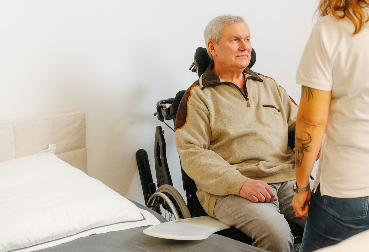 Älterer Mann sitzt im Rollstuhl neben dem Bett, eine Pflegefachkraft unterstützt ihn