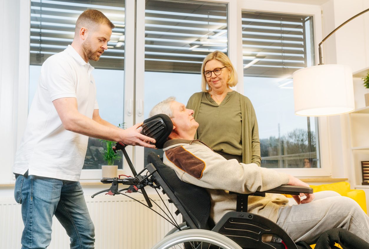 Carenetic-Mitarbeiter stellt die Kopfstütze eines Pflegerollstuhls ein, eine Angehörige schaut zu